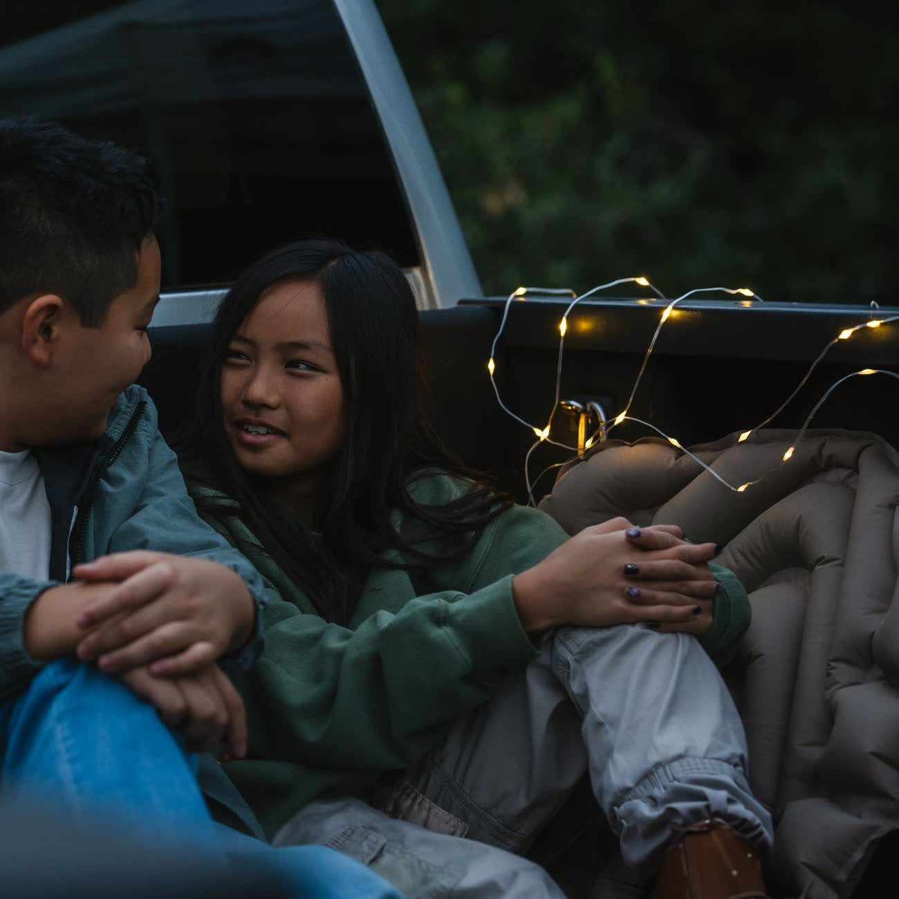 A man and a woman sitting in the bed of a pickup truck in the evening, illuminated by a LuminAID Solar String Light draped along the truck bed walls, creating a cozy ambiance.