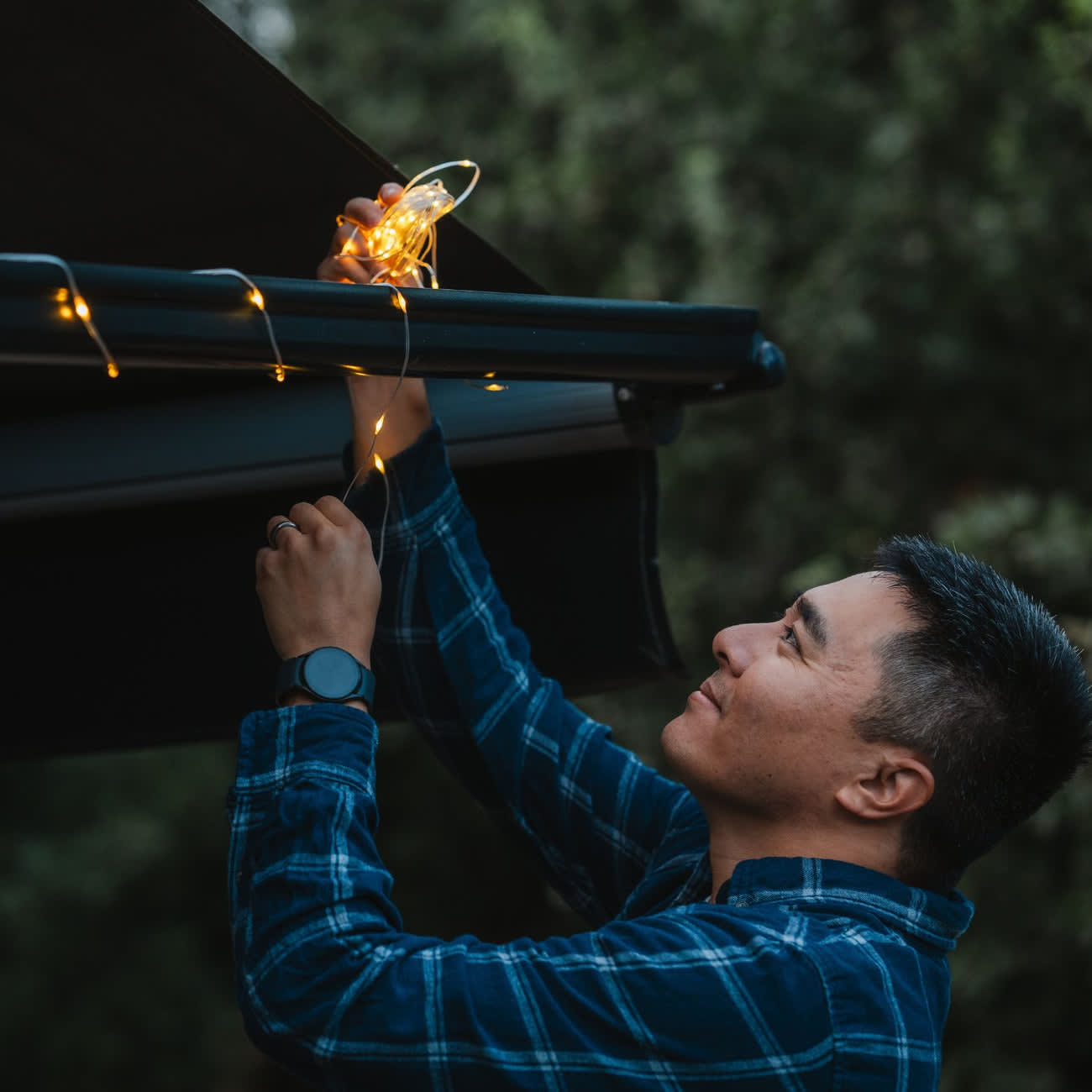 A man in a blue plaid shirt smiling as he hangs LuminAID Solar String Lights on the edge of a vehicle awning, with a soft warm glow from the lights against an outdoor backdrop.