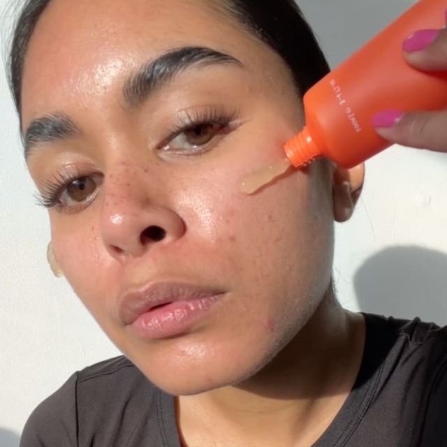 Person applying serum from an orange bottle onto cheek under sunlight.