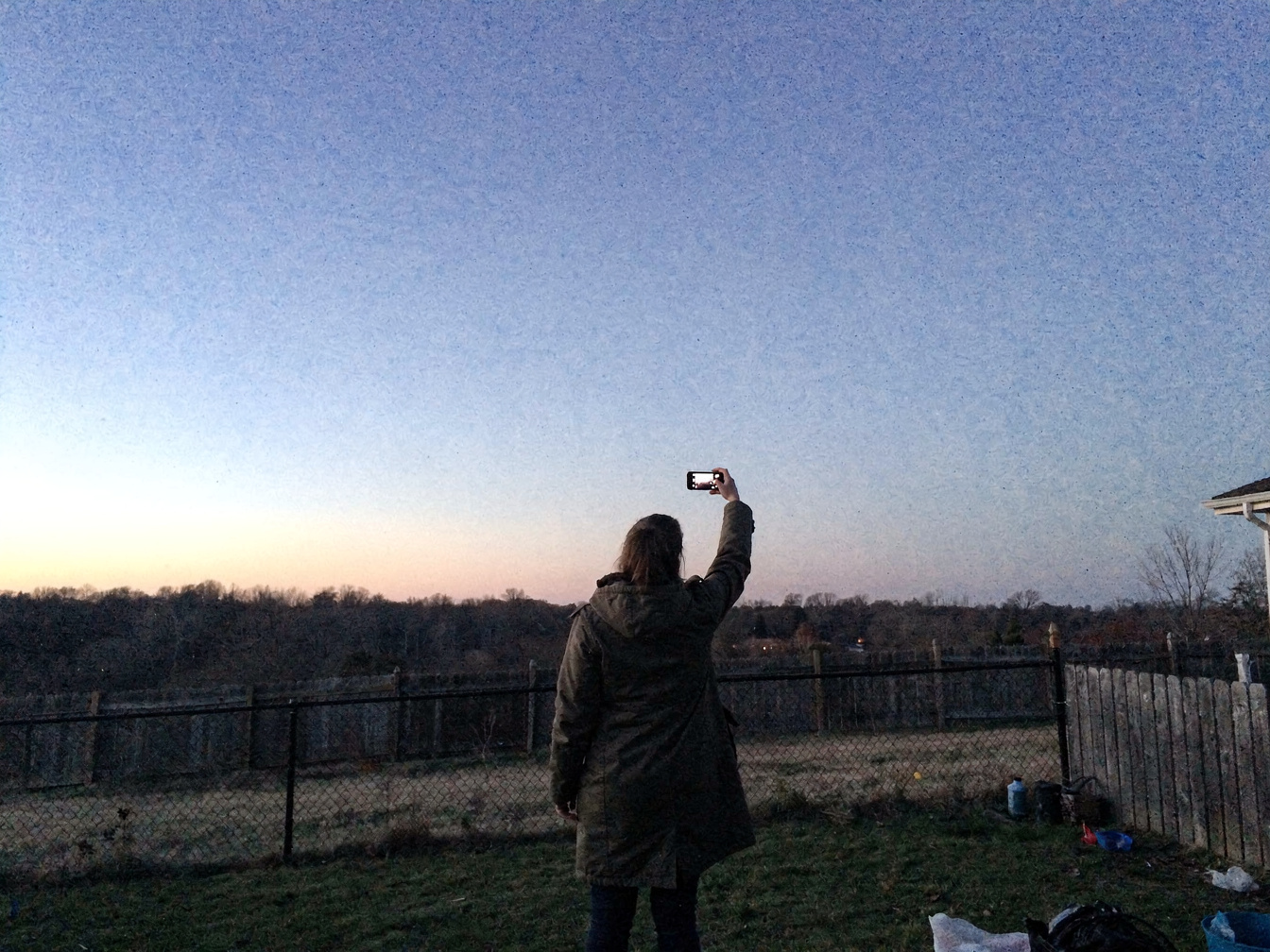 Seen from behind, a person alone in an open yard at dusk holding a phone up toward a clear sky.