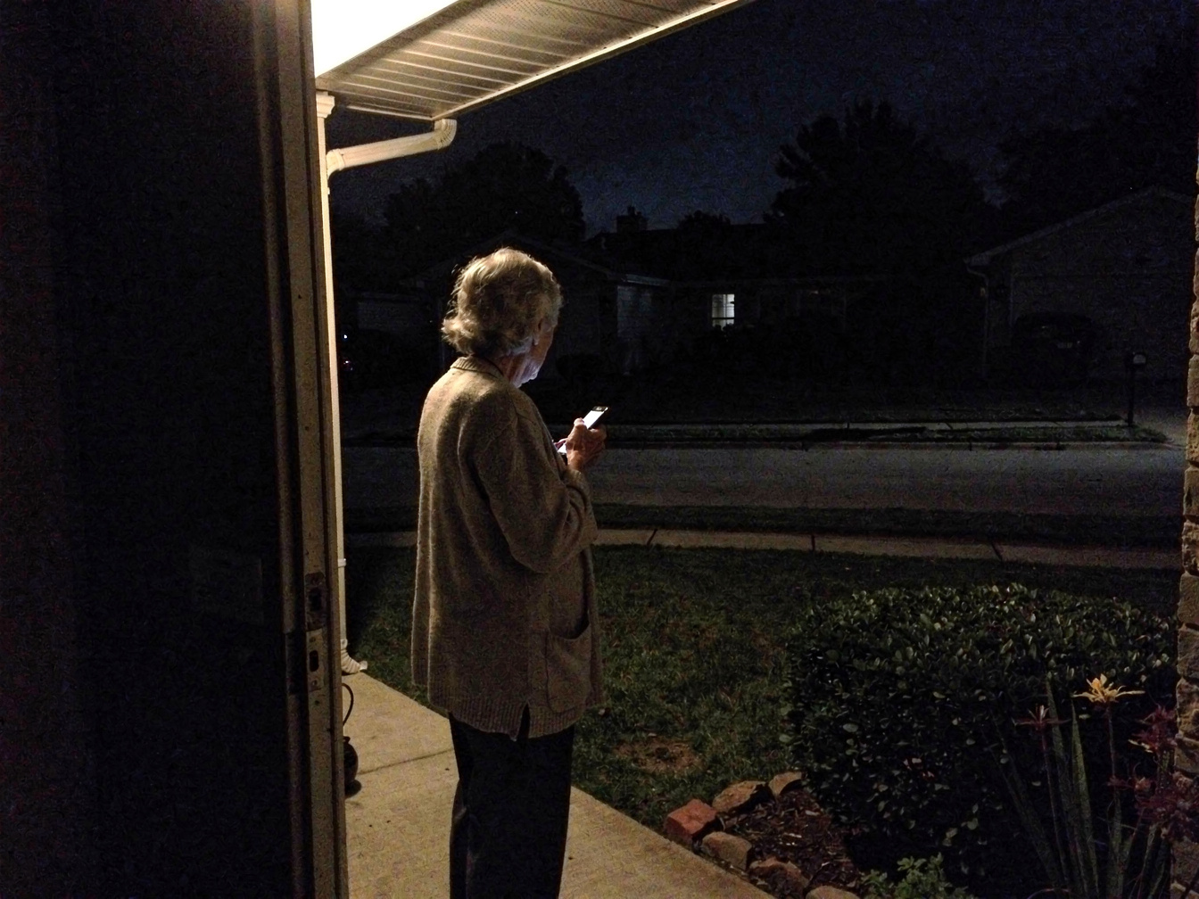 Illustration: an older person standing on a dark porch at night, checking a lit phone, the street behind them unlit.